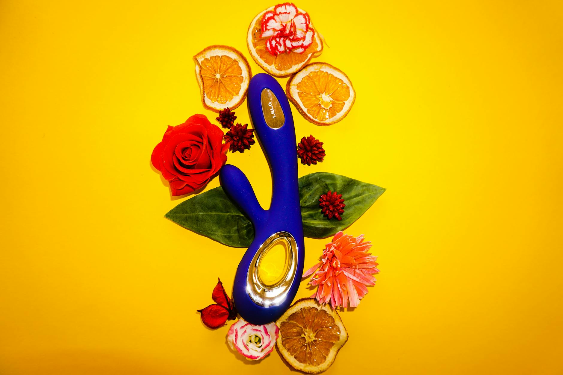A blue silicone clitoral vibrator surrounded by flowers and orange slices on a yellow background